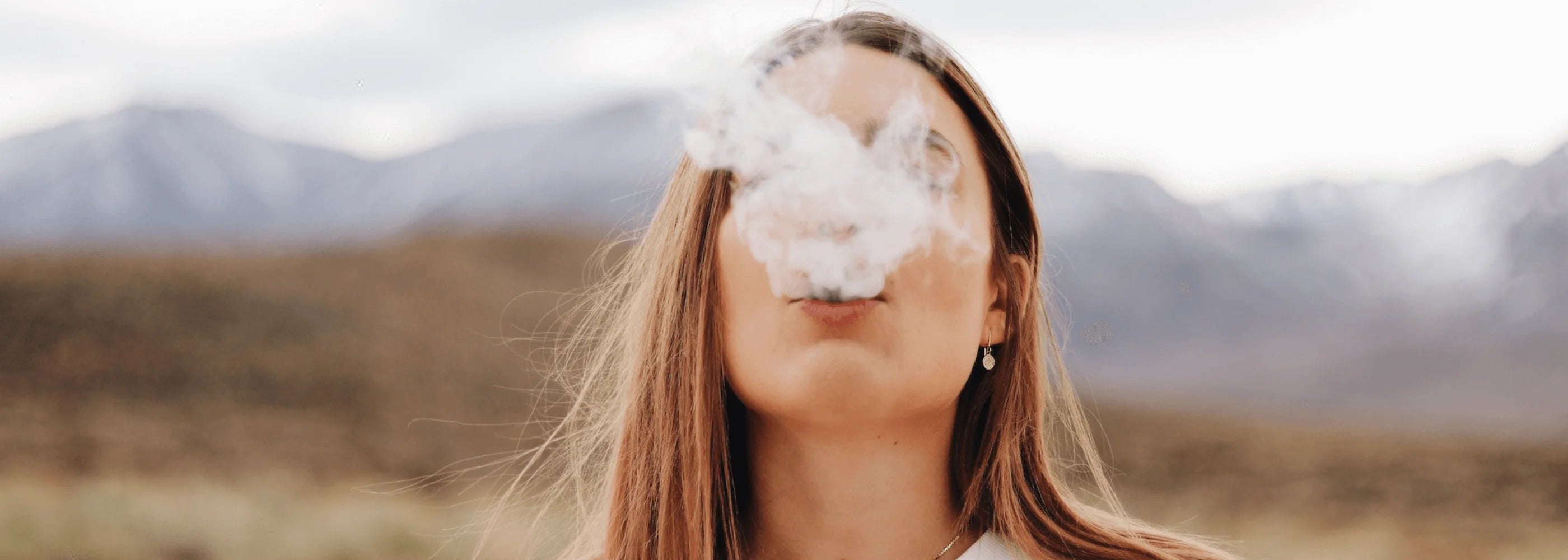 a blond girl exhaling a big puff with mountains in the background