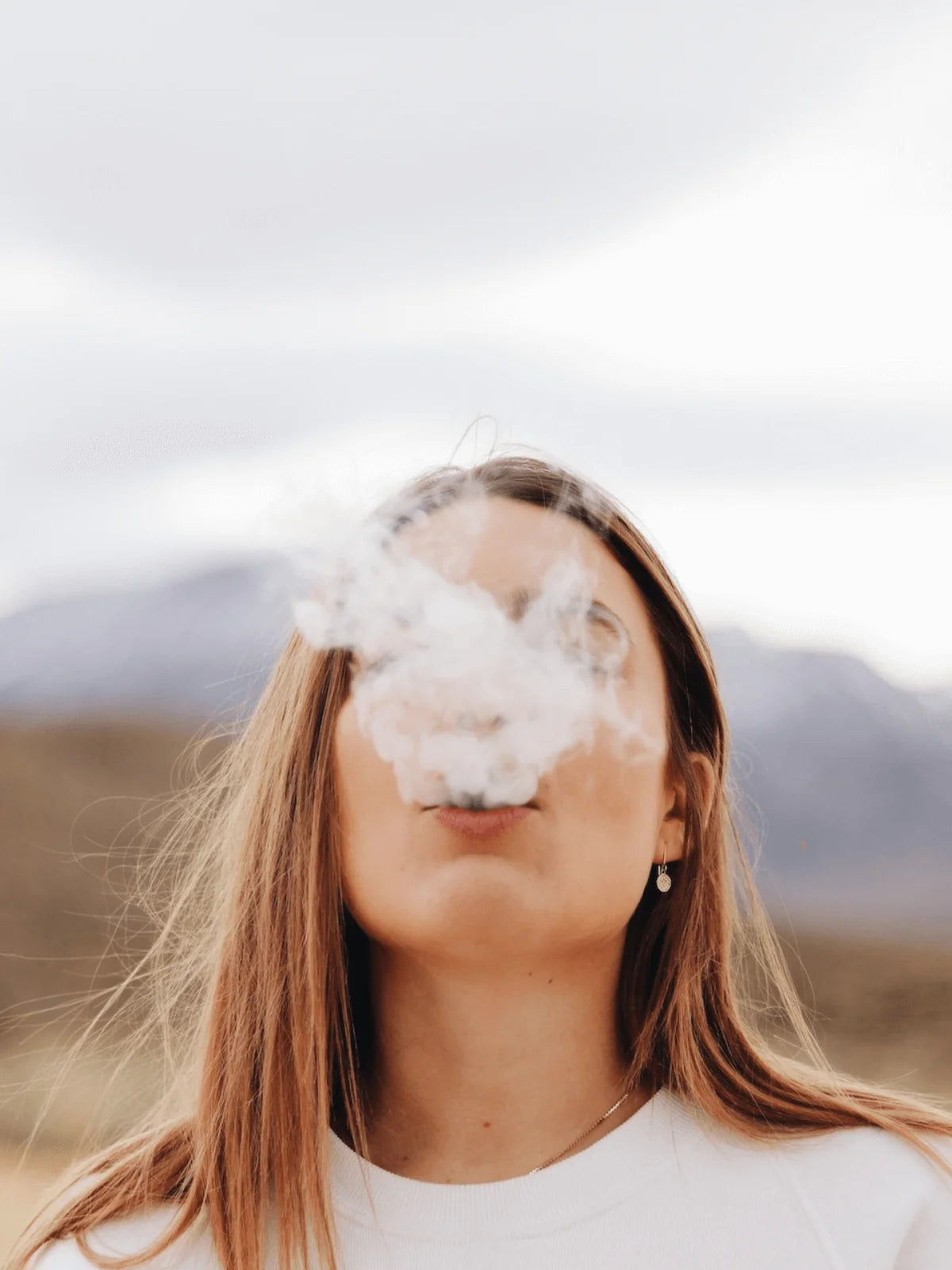 a blond girl exhaling a big puff with mountains in the background